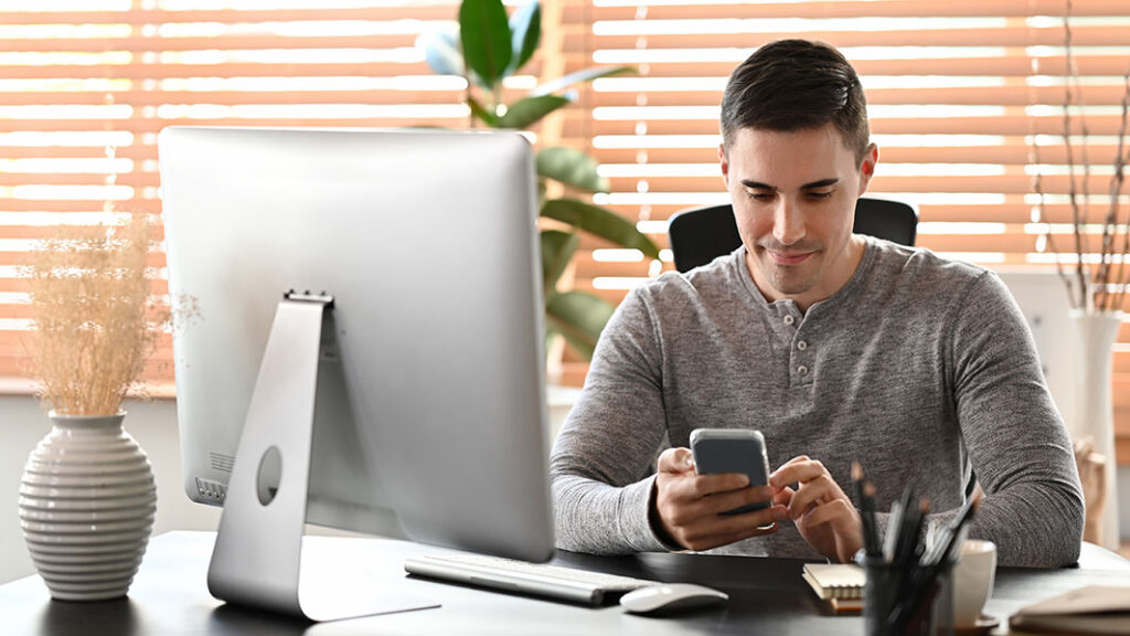 Person sitzt am Schreibtisch und tippt am Handy