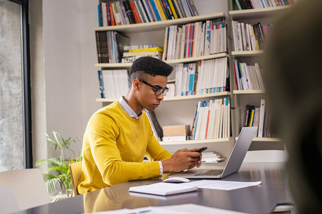 Junge sitzt vor Laptop mit Handy in der Hand