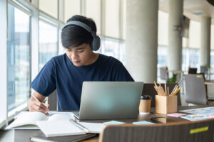 Junge sitzt vor Laptop und schreibt nebenher auf Papier